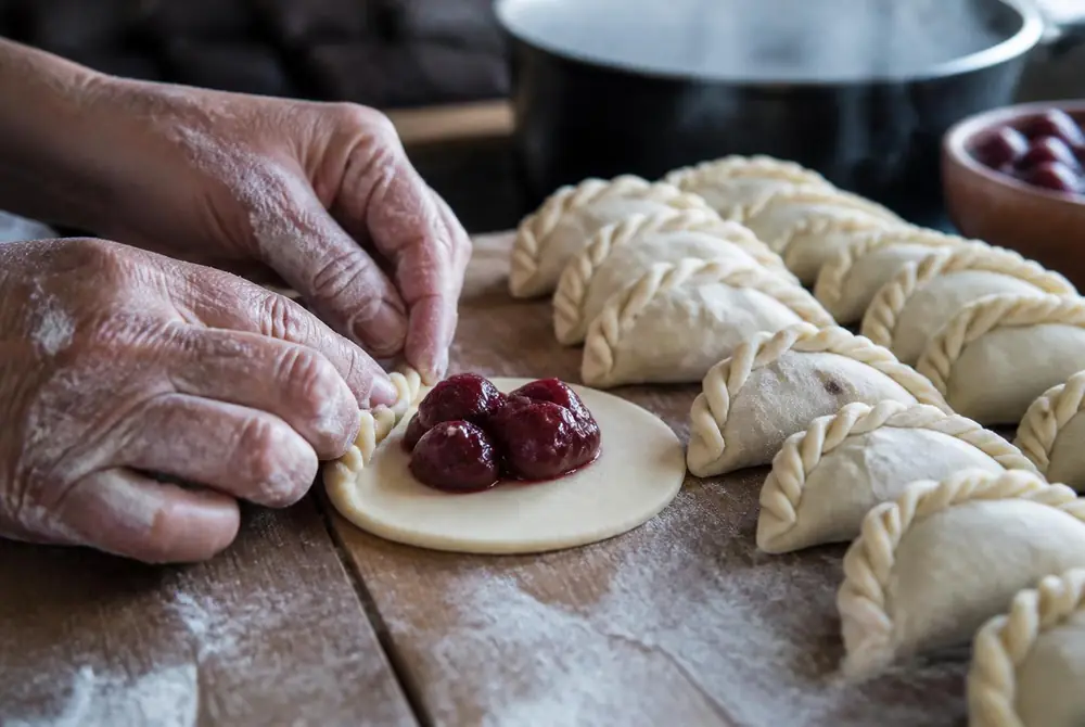 Hands crimping the edge of a cherry-filled dough round on a floured wooden board with rows of sealed varenikes beside a steaming pot