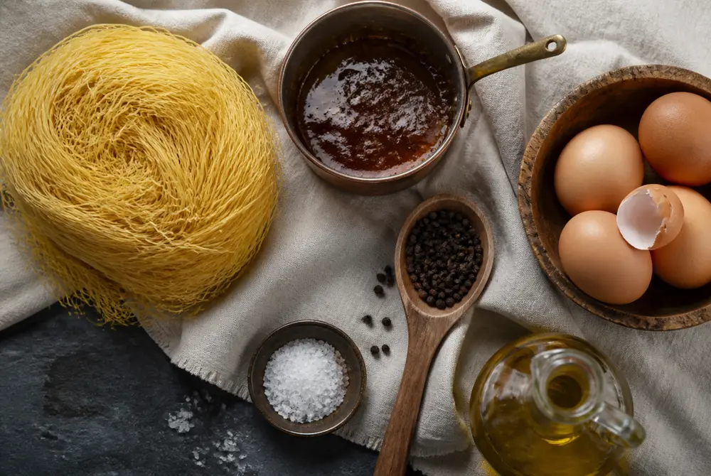 Overhead flat lay of Jerusalem kugel ingredients: thin angel hair noodles, dark caramel in a copper pot, brown eggs, black peppercorns, oil and coarse salt on linen