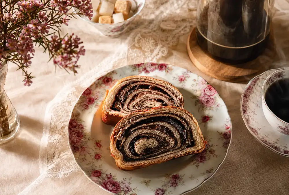 Two slices of chocolate kokosh cake showing dark cocoa spirals on a vintage floral plate beside a cup of black coffee on a warm linen tablecloth