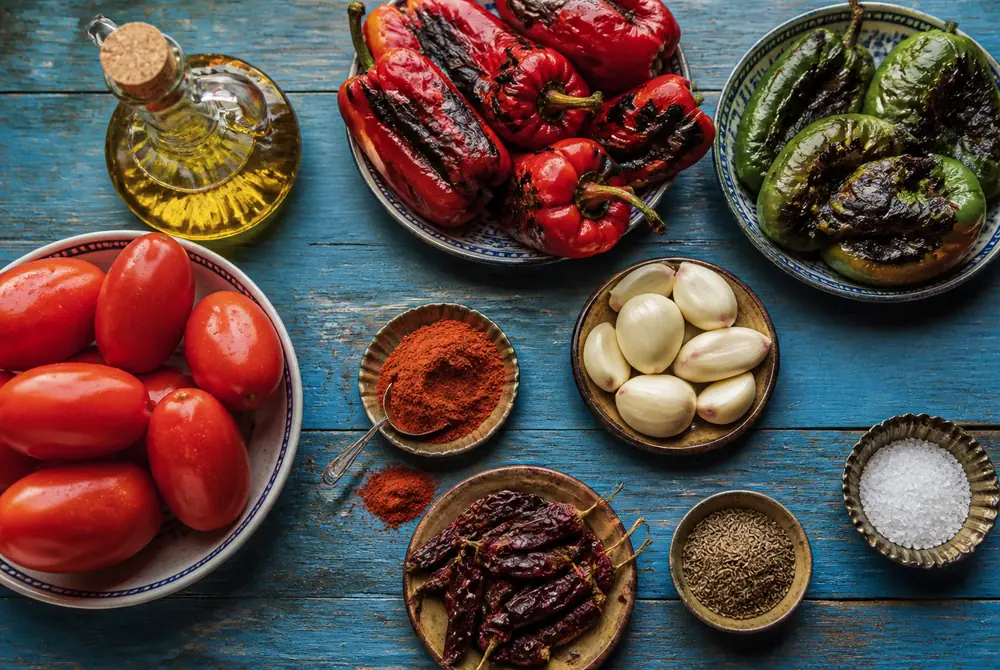 Overhead flat lay of matbucha ingredients including ripe plum tomatoes, charred red and green peppers, garlic cloves, sweet paprika, dried chilies, cumin and olive oil on a blue wooden table